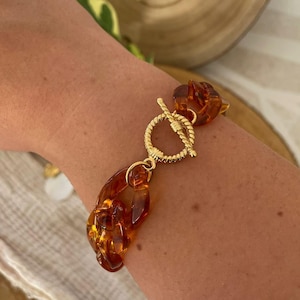 Peut inclure: Un bracelet chaîne doré avec des maillons de couleur ambre. Le bracelet est doté d'un fermoir à bascule et est porté au poignet. Les maillons ambre ont une finition brillante et le fermoir doré a un design texturé.