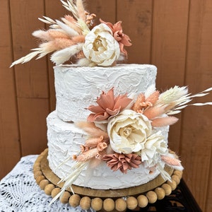 May include: Two-tiered white wedding cake decorated with dried flowers and pampas grass. The cake is on a wooden cake stand with a beaded edge.