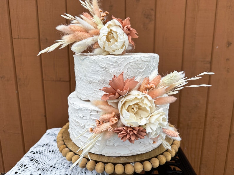 May include: Two-tiered white wedding cake decorated with dried flowers and pampas grass. The cake is on a wooden cake stand with a beaded edge.
