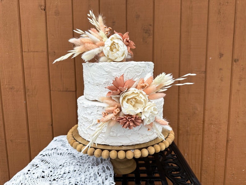 May include: Two-tiered white wedding cake decorated with dried flowers and pampas grass. The cake is on a wooden cake stand with a white lace tablecloth.