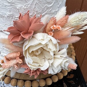May include: A white cake with a textured finish, decorated with a bouquet of dried flowers. The bouquet includes white and peach-colored pampas grass, white and peach-colored dried flowers, and a large white peony.