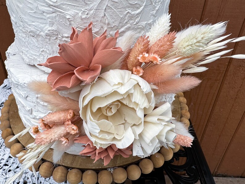 May include: A white cake with a textured finish, decorated with a bouquet of dried flowers. The bouquet includes white and peach-colored pampas grass, white and peach-colored dried flowers, and a large white peony.