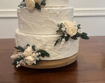 May include: A three-tiered white cake with textured frosting, adorned with cream-colored roses and greenery. The cake sits on a wooden cake stand, perfect for a wedding or special occasion.