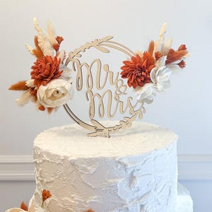 May include: A white wedding cake with a wooden "Mr & Mrs" topper decorated with dried flowers in shades of white, orange, and brown.