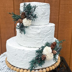 May include: A three-tiered white wedding cake with a rustic buttercream finish. The cake is decorated with sprigs of greenery, white flowers, and pine cones.