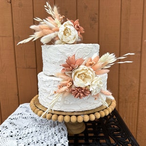 May include: Two-tiered white wedding cake decorated with dried flowers and pampas grass. The cake is on a wooden cake stand with a beaded edge.