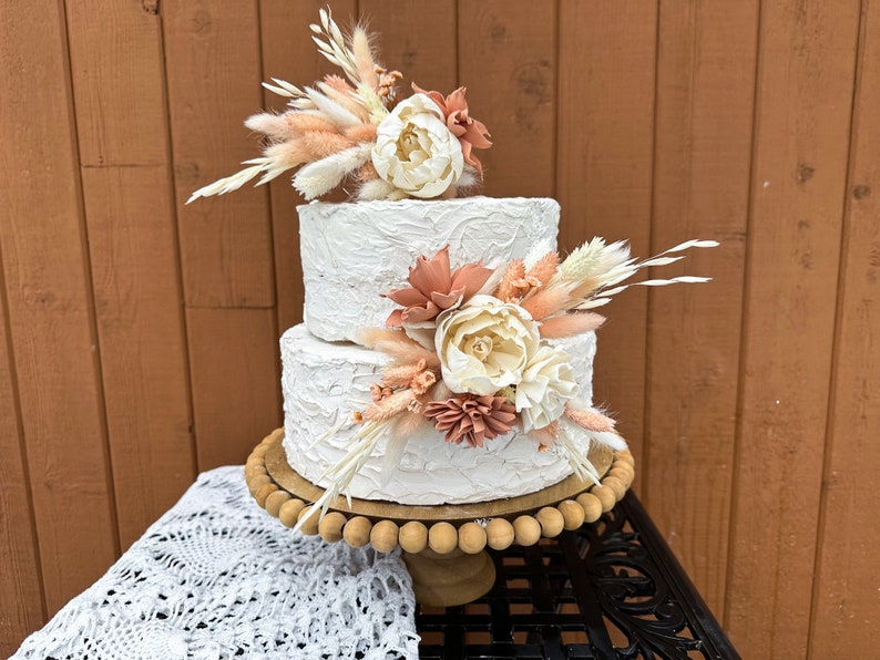 May include: Two-tiered white wedding cake decorated with dried flowers and pampas grass. The cake is on a wooden cake stand with a beaded edge.