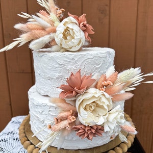 May include: Two-tiered white wedding cake decorated with dried flowers and pampas grass. The cake has a textured buttercream frosting and is topped with a cluster of white and peach-colored flowers.