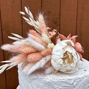 May include: A white cake with a pink and white dried flower arrangement on top. The arrangement includes pampas grass, dried peonies, and other dried flowers.