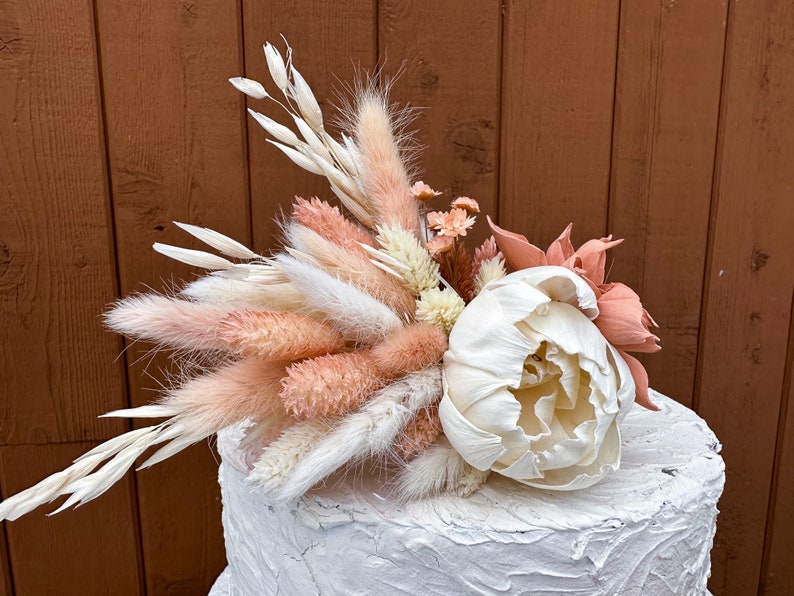 May include: A white cake with a pink and white dried flower arrangement on top. The arrangement includes pampas grass, dried peonies, and other dried flowers.