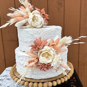 May include: Two-tiered white wedding cake decorated with dried flowers and pampas grass. The cake has a textured buttercream frosting and is displayed on a wooden cake stand.