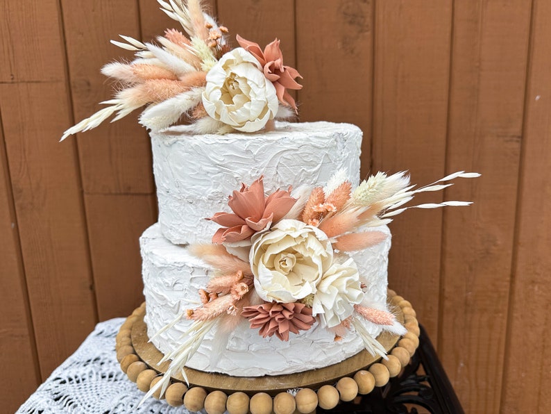 May include: Two-tiered white wedding cake decorated with dried flowers and pampas grass. The cake has a textured buttercream frosting and is displayed on a wooden cake stand.