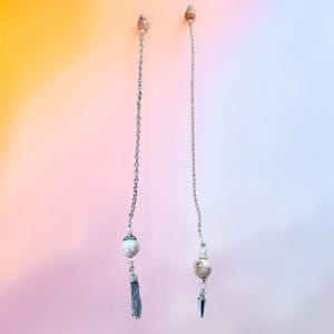 Puede incluir: Dos cadenas largas de tono plateado cuelgan verticalmente. Cada cadena presenta una pequeña cuenta redonda en la parte superior, una cuenta de piedra veteada más grande en el centro y un elemento decorativo de metal plateado, una borla y un cono.