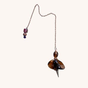 May include: A copper-toned metal chain features a brown tiger's eye gemstone pendant with a pointed silver-toned metal tip. The chain includes a small, dark purple patterned bead and a copper-toned spacer bead near the top end of the chain.