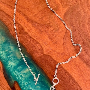 May include: A delicate silver necklace with a toggle clasp and a lobster clasp. The necklace is displayed on a wooden surface with a turquoise resin river design. The chain is made of small, interconnected links.