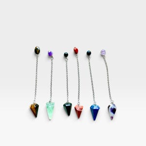 May include: Six pendulums featuring silver-toned metal chains, each topped with a small round gemstone bead and ending in a faceted, cone-shaped stone point. The stones include tiger's eye, green fluorite, black obsidian, red jasper, blue sodalite, and amethyst.