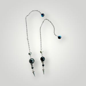 May include: Two silver chain earrings with dangling beads and spikes. Each earring features a chain with a dark blue bead at the top, followed by a series of smaller beads in black, white, and speckled patterns. The earrings end with a large, dark bead and a silver spike.