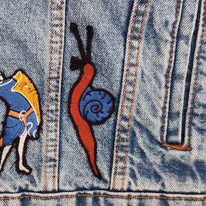 May include: Close-up of a light blue denim jacket with two embroidered patches. One patch depicts a medieval figure in blue and gold armour holding a staff. The other patch is a red snail with a blue shell and black antennae.