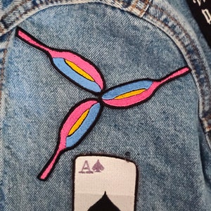 May include: A light blue denim jacket featuring several embroidered patches. Notable patches include a floral design in pink, blue, and yellow, a playing card with the Ace of Spades, and a black patch with white text: "We are the Daughters of the Witches You couldn't burn."