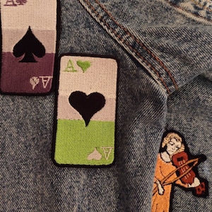 May include: A denim jacket with embroidered patches. The patches are of playing cards, a violin player, and a swirl design. The playing card patches are of the Ace of Spades and the Ace of Hearts.