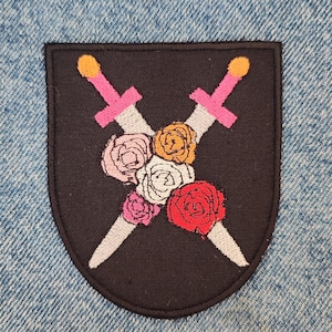 May include: Embroidered patch with a black shield design. Two swords cross over a bouquet of roses. The swords have pink handles and gold tips. The roses are red, pink, white, and orange.