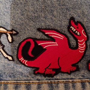 May include: Embroidered patches of a knight on horseback, a red dragon with white detailing, and a patterned lion, all sewn onto denim. The patches evoke a medieval or fantasy aesthetic, suitable for clothing or accessories.