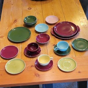 May include: A collection of colorful ceramic dishware, including plates, saucers, bowls, and teacups, in shades of green, pink, blue, and burgundy. The items are arranged on a wooden table.
