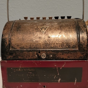 May include: A vintage, dark brown metal cash register with a red base. The register has a handle on top and a gold-colored label that reads "TODD VISIB" and "THE TODD COMPANY".
