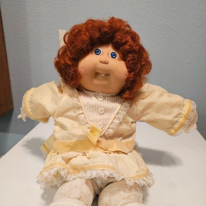 May include: A Cabbage Patch Kid doll with red curly hair, blue eyes, and a yellow and white dress. The doll is sitting on a white surface.