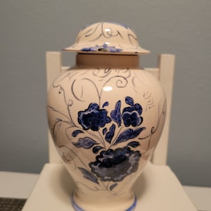 May include: A white ceramic vase with a blue and white floral design. The vase has a lid and a round body. The floral design features blue flowers and leaves on a white background.