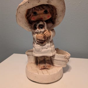 May include: A ceramic figurine of a girl wearing a large, wide-brimmed hat and a white coat, holding a small dog. The figurine has a light beige color with brown hair and boots. Two small buckets are at the girl's feet.