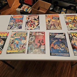 May include: A collection of vintage comic books, featuring titles like "Weird," "Human Fly," and "Thor." The covers display vibrant illustrations of superheroes, action scenes, and science fiction themes. The comics are arranged on a white surface.