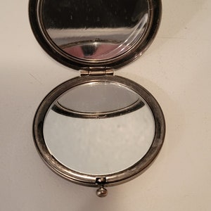 May include: A silver compact mirror with a round, hinged lid. The mirror is open, revealing a clear reflection.