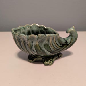 May include: A ceramic, shell-shaped bowl with a green and white marbled glaze. The bowl has a scalloped rim and a decorative, curved handle. It rests on a pedestal base with ornate detailing. The bowl is approximately 6 inches long.