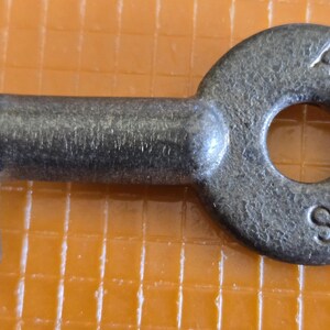 May include: A vintage metal key with a circular head and a rectangular bit. The key is a dark, aged gray color, with the letters "ATS RYS" stamped on the head. The key is lying on an orange, grid-patterned surface.
