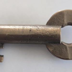 May include: A vintage brass key with a circular head and a cylindrical shaft. The head is stamped with the letters "NYCS". The key has a rectangular bit. The key is likely from a lock or a piece of machinery.