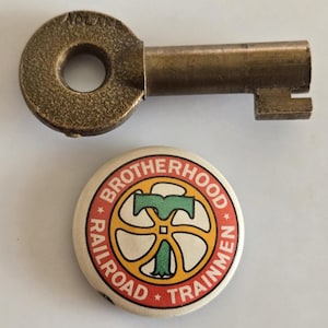 May include: A vintage brass key with the word "ADLAKE" stamped on the circular head, and a round button with the text "BROTHERHOOD RAILROAD TRAINMEN" encircling a logo with a green "T" and a yellow wheel design.