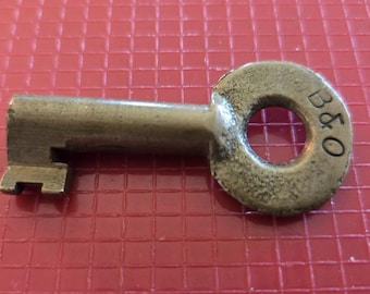 May include: A silver-toned metal key with a round head. The head has a hole in the center and the letters "B&O" engraved on the side.