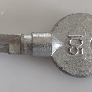 May include: A silver-colored key with a circular head and a rectangular bit. The head has a hole and the number "105" stamped on it. The key is likely for a lock or other mechanical device.