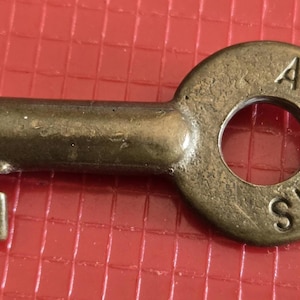 May include: A brass-colored metal key with a circular bow featuring the stamped text AT-SF RYS. The key has a cylindrical shank and a notched bit at the end. The metal surface shows signs of age and wear with a dull, slightly tarnished finish.