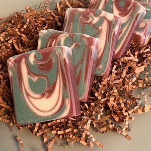 May include: Five handmade soap bars with a swirl pattern of green, white, and red. The bars are arranged in a pile on brown shredded paper.