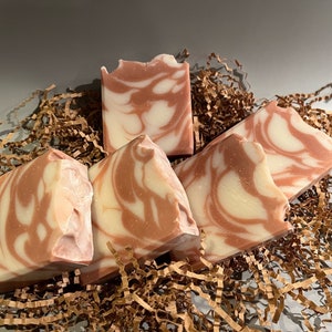 May include: Four bars of handmade soap with a swirl pattern of pink and white. The soap is sitting on a bed of brown shredded paper.