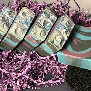 May include: Four bars of handmade soap with a swirling blue, pink, and gray pattern. The soap is displayed on a bed of purple shredded paper.