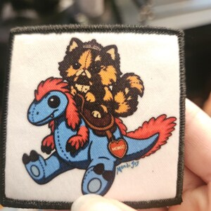 May include: A square fabric patch with a black stitched border featuring a cartoon illustration of a fluffy brown and black cat riding a blue dinosaur with red accents. A brown saddle with a red heart-shaped tag is visible on the dinosaur's back.