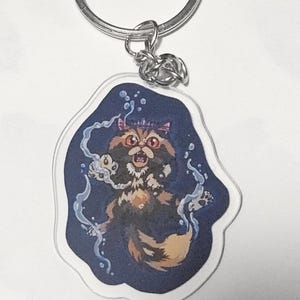 May include: A clear acrylic keychain featuring a printed illustration of a fluffy, multi-colored cat with large orange eyes and a crown, surrounded by swirling blue water effects and bubbles, attached to a silver-toned metal split ring.