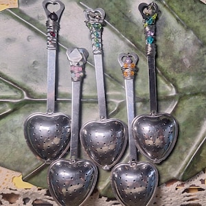 May include: Six silver heart-shaped tea infusers with different colored beads and charms on the handles. The infusers are on a green and brown background.