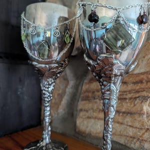 May include: Two silver-toned metal goblets featuring intricate embossed grape vine and leaf detailing on the stems and bases. Each goblet is adorned with a delicate silver chain draped around the rim, featuring small dangling charms and wire-wrapped stones.
