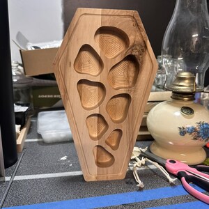 Puede incluir: Una bandeja de madera en forma de ataúd con seis compartimentos tallados. La bandeja está hecha de madera clara, con un patrón de vetas naturales. Los compartimentos están diseñados con un interior texturizado y en capas.