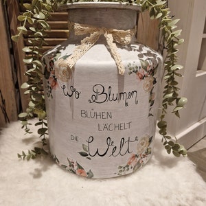 Könnte beinhalten: Eine weiße Keramik-Milchkanne mit Blumenmuster und dem Text "Wo Blumen Blühen Lächelt Die Welt". Die Kanne ist mit einem Spitzenband verziert und hat einen Zweig Grün auf der Oberseite.
