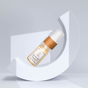 Puede incluir: Un pequeño frasco de vidrio ámbar con una tapa de madera y un dispensador de bomba blanco. El frasco está etiquetado como "Eye Like Steel" y contiene un suero para los ojos. El frasco está sentado sobre una superficie blanca curva.